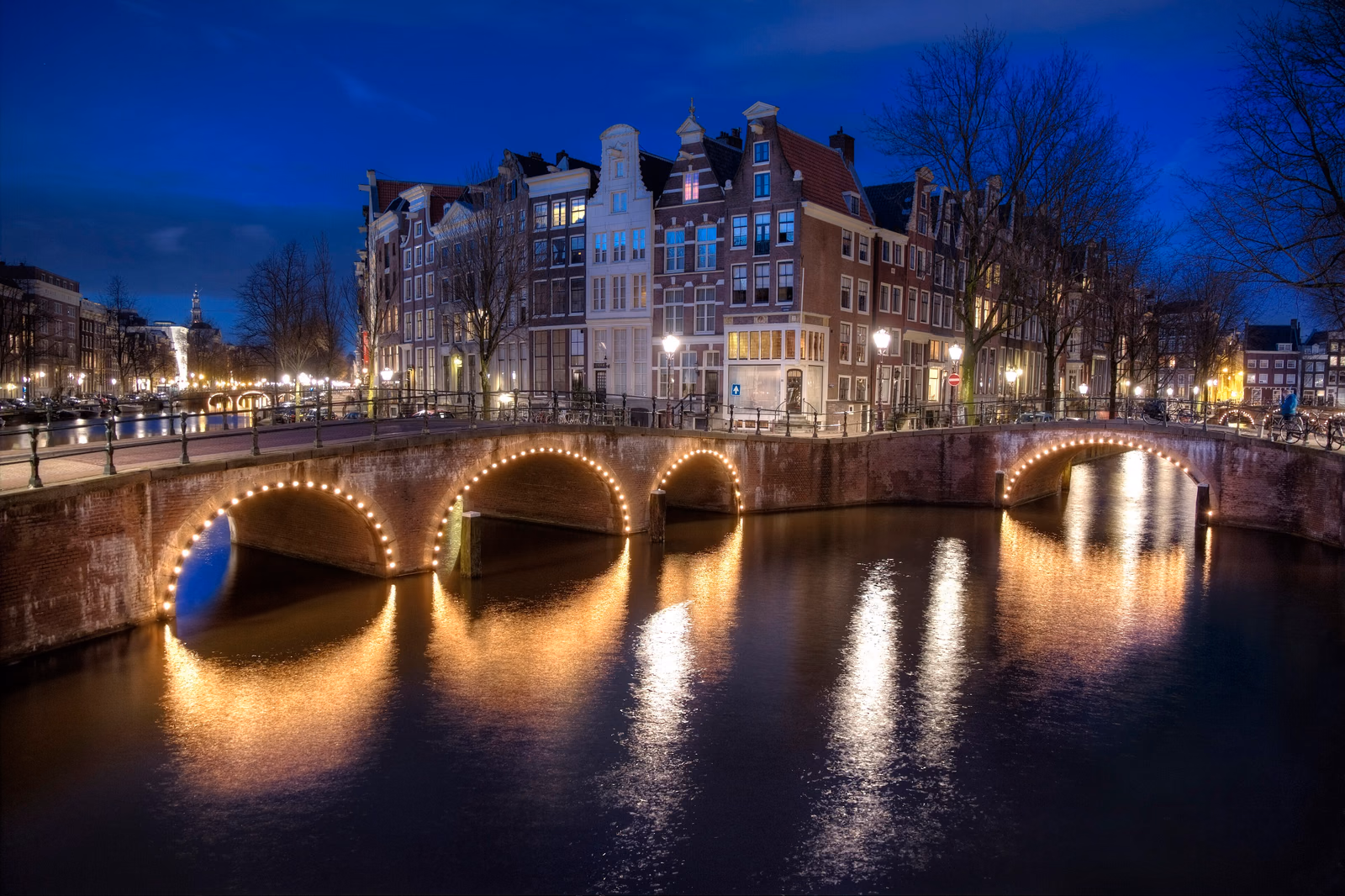 Beleuchtete Brückenbögen an einer Amsterdamer Gracht mit Giebelhäusern und kahlen Bäumen zur blauen Stunde
