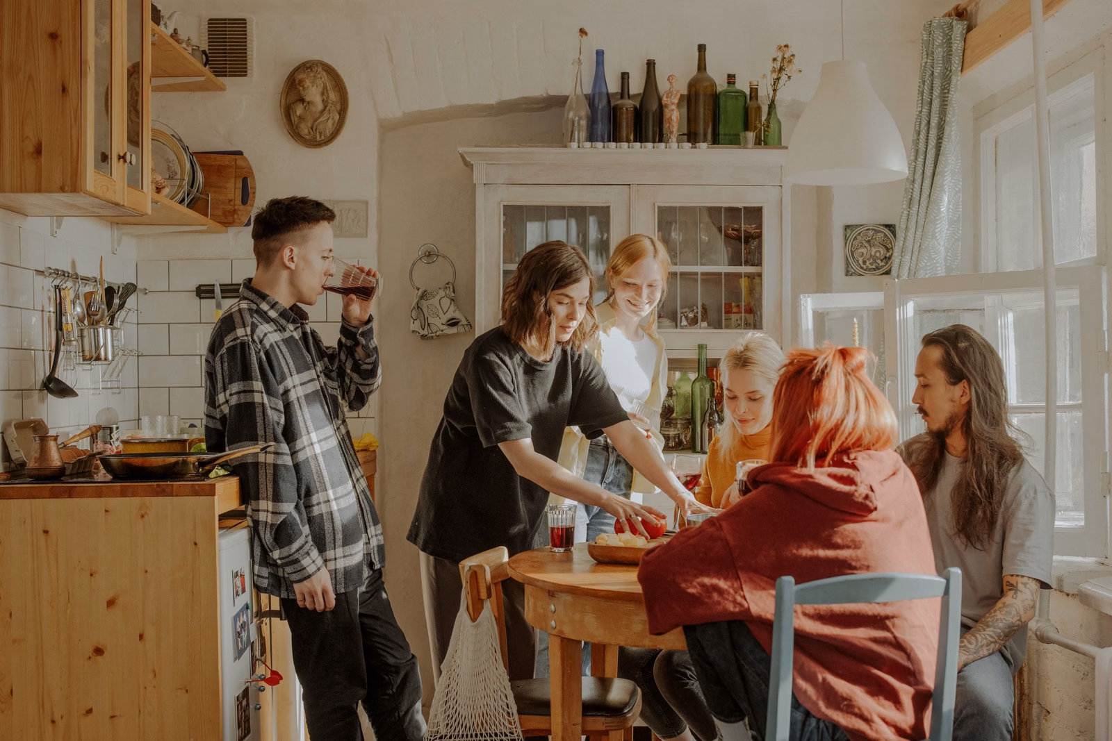 Sechs Freunde in einer rustikalen Küche um einen runden Holztisch, eine Frau stellt eine Schale mit Essen ab